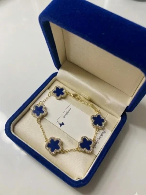Guldarmband med blå klöverhängen 18K pläterad - Snyggt armband i guldton med fem blå klöverformade hängen, kantade av små gnistrande stenar. Länken är tunn och justerbar för perfekt passform. Perfekt accessoar för att lyfta din outfit med en färgklick och elegant design.
