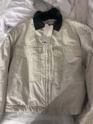 Beige overshirt från F3 Sister - Stilren beige overshirt från F3 Sister med svart krage och två stora bröstfickor. Overshirten har rak passform och är tillverkad i ett slitstarkt bomullstyg. Perfekt att bära öppen eller stängd för en avslappnad look.