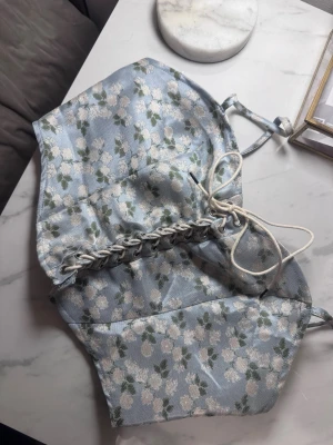 Blommig korsettopp från Cider - Supersöt ljusblå korsettopp från Cider med vita blommor och gröna blad. Toppen har snörning framtill och tunna axelband. Materialet är glansigt och känns som satin, perfekt för att lyfta vilken outfit som helst.