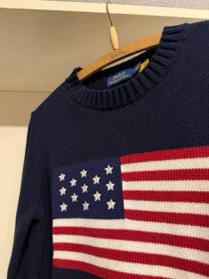USA flagga stickad Ralph Lauren - Mörkblå stickad tröja från Polo Ralph Lauren med amerikansk flagga i rött, vitt och blått på bröstet samt RL-broderi. Tröjan har rund halsringning och ribbade muddar vid ärmslut och nederkant. Perfekt för dig som gillar klassisk preppy stil. Passar xs/s