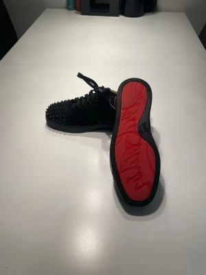 Svarta Christian Louboutin sneakers - Säljer ett par svarta sneakers från Christian Louboutin med ikoniska röda sulor och svarta nitar över hela tåpartiet. Skorna har snörning och är tillverkade i mocka och läder för en lyxig känsla. Perfekta för dig som vill sticka ut med en edgy och exklusiv stil.