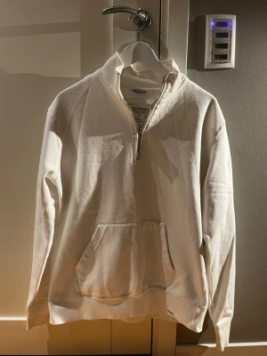 Vit halfzip hoodie från pull&bear - Vit hoodie från pull&bear med halv dragkedja framtill och stor magficka. Tillverkad i mjukt bomullsmaterial och har ribbade muddar vid ärmslut och nederkant. Klassisk och enkel design utan tryck eller mönster, perfekt för en clean look.