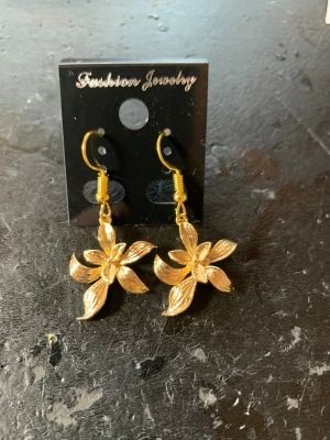 Guldiga blommiga hängörhängen - Snygga örhängen i guldfärgad metall med blomformad design. Varje blomma har fem detaljerade kronblad och örhängena har krokar i matchande färg. Perfekta för dig som vill ha en unik och trendig accessoar med blommig touch.