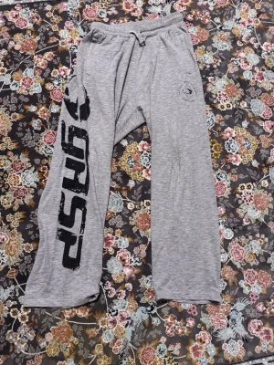 Gråa mjukisbyxor från GASP - Snygga grå mjukisbyxor från GASP med stort svart tryck längs ena benet och dragsko i midjan. Byxorna har avslappnad passform och är tillverkade i mjukt bomullsmaterial. Perfekta för chill eller gymmet.