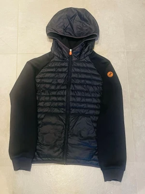 Svart hybridjacka från Save The Duck - Snygg svart hybridjacka från Save The Duck med quiltad front och huva. Jackan har dragkedja framtill, ribbade muddar och ett orange logomärke på ärmen. Perfekt för dig som vill ha en stilren och funktionell jacka.