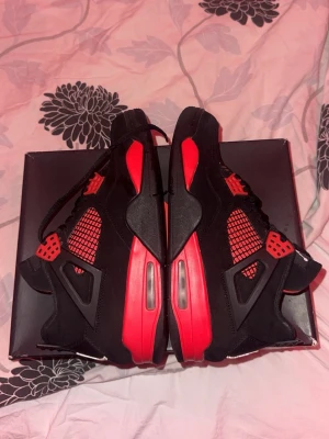 Jordan 4 red Thunder ÄKTA Dom är begagnade - Grymma Air Jordan 4 sneakers i svart och rött med meshpaneler och detaljer i plast. Skorna har en chunky sula med synlig Air-enhet och coola röda accenter på sidorna och sulan. Ovandelen är i mocka och textil, perfekt för dig som gillar streetstyle.