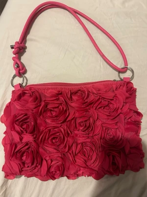 Rosa handväska med rosdetaljer - Snygg rosa handväska med 3D-rosor över hela framsidan. Väskan har ett axelband i matchande färg och stängs med dragkedja. Perfekt för dig som vill sticka ut med en unik accessoar. Materialet är tyg med textilrosor och detaljer i metall.