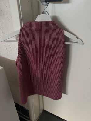 Ribbad rosa ärmlös topp glittrig - Snygg ribbad topp i vinröd färg med hög hals och ärmlös design. Perfekt att styla med jeans eller kjol för en trendig look. Mjuk och stretchig kvalitet som sitter skönt på kroppen.