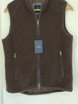 Brun pileväst från Hansen & Jacob - Mysig brun pileväst från Hansen & Jacob med hög krage och två dragkedjeförsedda fickor framtill. Västen har hel dragkedja och är gjord i mjukt teddyfleece-material som ger en avslappnad och trendig look.