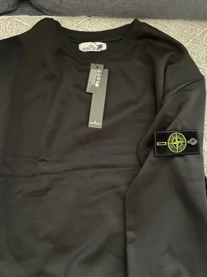 Svart sweatshirt från Stone Island - Säljer en svart sweatshirt från Stone Island med klassisk rund hals och ribbade muddar. Tröjan har den ikoniska Stone Island-loggan på vänster ärm och är tillverkad i mjukt bomullsmaterial. Perfekt för en stilren och avslappnad look.