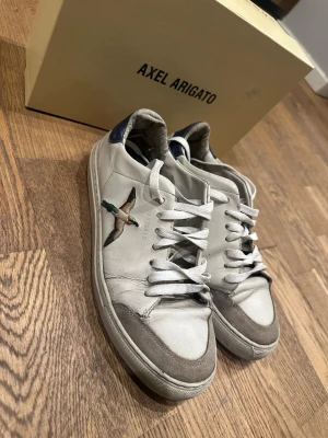 Vita Axel Arigato sneakers med fågel - Snygga vita sneakers från Axel Arigato med broderad fågel på sidan. Skorna har grå mockadetaljer vid tån, vita skosnören och blå hälparti. Tillverkade i läder och mocka för en clean och stilren look. Perfekta för dig som gillar streetstyle och unika detaljer.