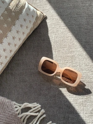 Sommriga solglasögon från Carin Wester - Supersnygga solglasögon med bred, ljusbeige båge och UV-skydd. Trendig design med markerad ram och diskret logga. Perfekta för dig som vill ha en stilen och modern look i solen. 