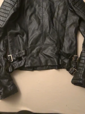 Svart biker skinnjacka med spännen - Snygg svart skinnjacka i bikerstil med dubbla spännen i midjan och dragkedjor framtill. Jackan har quiltade detaljer på axlarna och en kort, figurnära passform som ger en cool look. Perfekt för dig som vill ha en edgy stil.