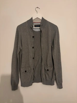 Grå cardigan från Massimo Dutti - Stilren grå cardigan från Massimo Dutti med knäppning framtill och två fickor med knapp. Tröjan har ribbade muddar vid ärmslut och nederkant samt en klassisk rund halsringning. Perfekt för lager på lager och enkel att matcha.