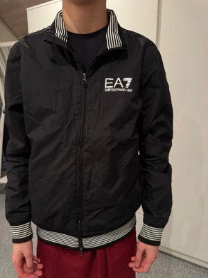 Svart bomberjacka EA7 Emporio Armani - Svart bomberjacka från EA7 Emporio Armani med vit logga på bröstet. Jackan har dragkedja framtill och ribbade muddar med svartvita ränder vid ärmslut, krage och nederkant. Snygg, sportig look med klassiska detaljer.