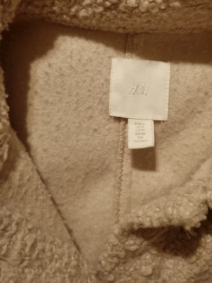 Beige fleece overshirt från H&M - Mysig beige overshirt från H&M i fleece med knappar framtill och bröstficka. Perfekt för lager på lager och har en avslappnad passform med klassisk krage. Materialet är mjukt och fluffigt, vilket ger en trendig look.