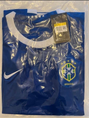 Brasilien 2006 fotbollströja. Helt ny! - Helt ny! Snygg blå fotbollströja från Nike med Brasilien CBF-emblem på bröstet och vita detaljer. Tröjan har rund halsringning med vit kant och flera stjärnor ovanför emblemet. Perfekt för dig som älskar fotboll och vill stötta Brasilien.