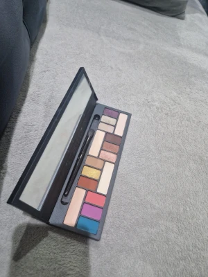 Smashbox ögonskuggspalett med spegel - Färgstark ögonskuggspalett från Smashbox med 12 nyanser i både matta och skimrande toner. Innehåller färger som blå, rosa, guld, lila, beige och brunt. Paletten har en stor spegel och medföljande applikator. Perfekt för kreativa sminkningar.