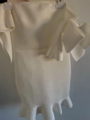 Vit kort klänning med volang och rosett - Superfin vit klänning med volangdetaljer och en stor rosett på axeln. Klänningen har en figurnära passform och är kort i modellen, perfekt för dig som vill sticka ut. Materialet känns slätt och har en lätt glans.