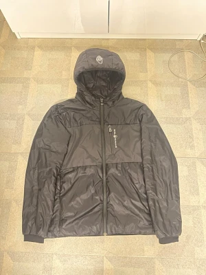 Svart pufferjacka från Sail Racing - Snygg svart pufferjacka från Sail Racing med huva och dragkedja framtill. Jackan har en bröstficka med logga och en broderad detalj på huvan. Funkar toppen som vindjacka i både vår,höst och sommar