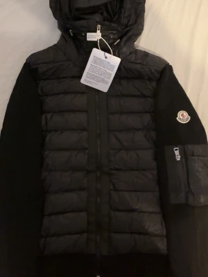 Svart dunväst från Moncler med huva - Snygg svart dunväst från Moncler med huva och dragkedja framtill. Västen har quiltad design, Moncler-logga på ärmen och en praktisk ficka med dragkedja. Insidan har ett unikt Moncler-tryck. Perfekt för lager-på-lager och streetstyle.