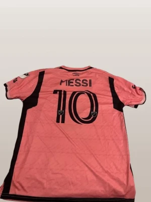 Rosa Inter Miami Messi matchtröja - Säljer en rosa Inter Miami matchtröja från Adidas med Messi och nummer 10 på ryggen. Tröjan har svarta detaljer längs sidorna och axlarna samt klubbmärke och MLS-logga. Perfekt för dig som älskar fotboll och vill stötta Messi.