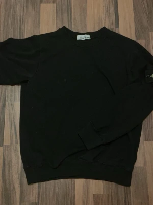 Svart sweatshirt från Stone Island - Snygg svart sweatshirt från Stone Island med klassisk rund halsringning och ribbade muddar. Tröjan har den ikoniska Stone Island-loggan på ärmen och är tillverkad i mjukt bomullsmaterial. Perfekt för en clean och stilren look.
