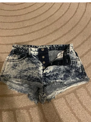 Acid wash denimshorts med låg midja - Säljer ett par snygga acid wash denimshorts med låg midja. Shortsen har fem fickor, bälteshällor och råa kanter nedtill för en edgy look. Perfekta till sommaren och ger en cool vintage vibe.