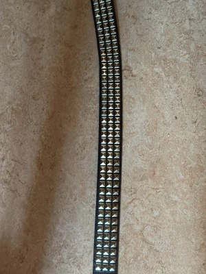 Svart nitar skärp med silverdetaljer - Snyggt svart skärp med silverfärgade fyrkantiga nitar längs hela bandet. Skärpet har en klassisk metallspänne och är perfekt för att ge din outfit en edgy vibe. Tillverkat i skinnimitation.