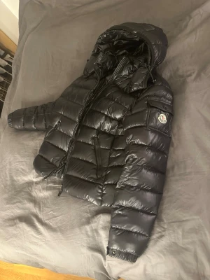 Svart dunjacka från Moncler - Snygg svart dunjacka från Moncler med glansig finish, huva och dragkedja framtill. Jackan har Moncler-logga på ärmen och praktiska fickor med dragkedja. Perfekt för kalla vinterdagar och riktigt stilren look.
