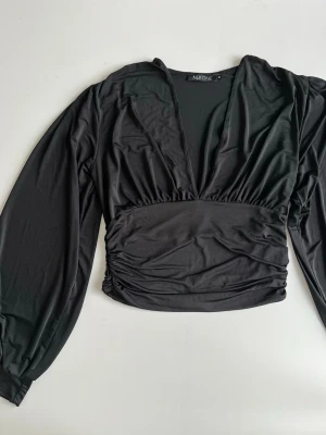 Svart croppad topp från NLY ONE - Svart croppad topp från Nelly med djup v-ringning och lite puffigare ärmar. Rynkade detaljer vid midjan mycket stretchig!! Lika snygg till kostymbyxor som jeans. Tunn och härlig till vår/sommarkvällar.