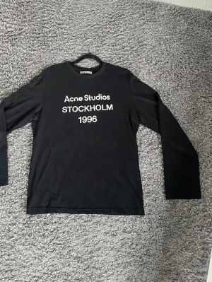 Acne Studios sthokholm 1996 - Svart långärmad topp från Acne Studios med vit text 'Acne Studios STOCKHOLM 1996' på bröstet. Klassisk rund halsringning och rak passform. Perfekt för dig som gillar stilrena och trendiga plagg med tydlig logga.