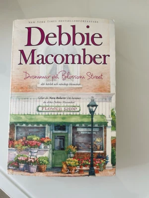 Drömmar på Blossom Street - En mysig roman av Debbie Macomber med ett charmigt omslag som visar en blomsterbutik. Boken är tryckt på papper och har en färgglad framsida med gröna, rosa och vita toner. Perfekt för dig som gillar feelgood och romantik.