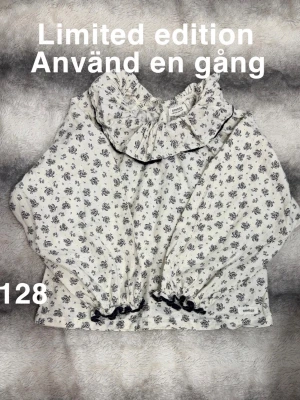 Blommig volangblus från Newbie  - Supersöt blus från Newbie med svart blommönster på vit botten. Volangkrage med svart kant och volangdetaljer vid ärmslut och midja. Långärmad modell. Perfekt för dig som gillar unika plagg med fina detaljer.🌸 Använd en gång. Limited edition. Storlek 128