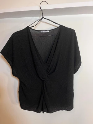 Svart omlottopp från Zara - Svart omlottopp från Zara med djup v-ringning och twistad detalj framtill. Toppen har korta ärmar. 