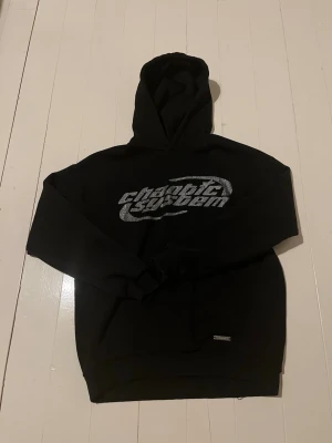 Svart rhinestone hoodie - Snygg svart hoodie med rhinestone logga på bröstet. Klassisk passform med huva och ribbade muddar. Perfekt för dig som gillar streetwear och vill ha en stilren och basic look. 