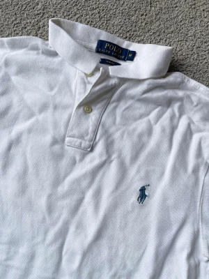 Vit pikétröja från Polo Ralph Lauren, storlek S - Vit pikétröja från Polo Ralph Lauren med klassisk krage, två knappar vid halsen och blå broderad logga på bröstet. Tillverkad i mjuk bomull och har korta ärmar för en clean och tidlös stil. 