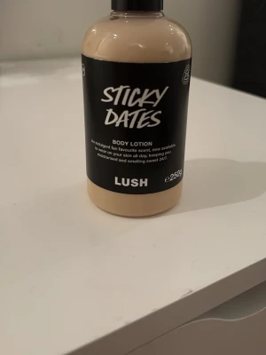 LUSH Sticky Dates Body Lotion 250g - Sticky Dates Body Lotion från LUSH i en beige plastflaska med svart etikett. Innehåller 250g återfuktande lotion med söt doft, perfekt för att hålla huden mjuk och väldoftande hela dagen. Snygg och enkel design som passar på badrumshyllan. Endast testad en gång!