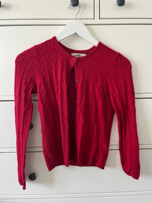 Röd kabelstickad kofta - Snygg röd kabelstickad kofta med rund halsringning och knappar framtill. Långärmad modell med ribbade muddar vid ärmslut och nederkant. Storlek 146/152 men passar xs/s skulle jag säga. 