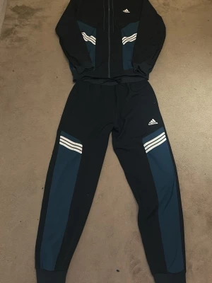 Adidas mörkblå träningsset - Snyggt träningsset från Adidas i mörkblått med blå detaljer och klassiska vita ränder. Setet består av en zip-hoodie med dragsko och matchande byxor med resår i midjan. Perfekt för gymmet eller chill. Materialet är mjukt och stretchigt för maximal komfort.