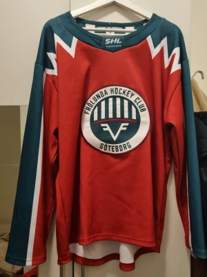 Frölunda HC hockeytröja röd/grön - Säljer en snygg Frölunda Hockey Club matchtröja i rött och grönt med klubbens logga på bröstet. Tröjan har långa ärmar, vita detaljer och är tillverkad i ett lätt och slitstarkt material. Perfekt för hockeyfans som vill stötta sitt lag på match eller träning.