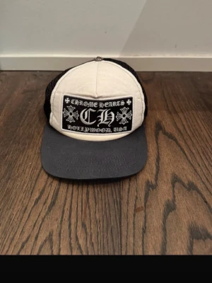 Chrome Hearts keps - Säljer en stilren truckerkeps från Chrome Hearts i svart och vitt. Kepsen har ett broderat märke framtill med text och blommotiv, svart mesh baktill och en metallknapp med gravyr på toppen. Skärmen är svart och framsidan är vit i tyg.