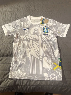 Jesus Brasilien fotbollströja Nike - Snygg vit fotbollströja från Nike med Brasilien-emblem och detaljerad grafik av Kristusstatyn och brasilianska motiv. Tröjan har blå och gula inslag, rund hals och är tillverkad i Dri-FIT material som andas. Perfekt för dig som vill sticka ut på planen.
