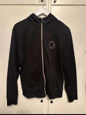 Svart zip-hoodie från BOSS - Svart hoodie med dragkedja från BOSS. Klassisk modell med huva och dragsko, samt fickor framtill. Liten BOSS-logga i vitt på bröstet. Perfekt för en chill och stilren look.