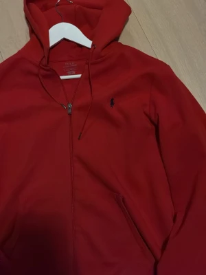 Röd zip up från Polo Ralph Lauren - Snygg röd hoodie från Polo Ralph Lauren med dragkedja framtill och klassisk huva med dragsko. Ikoniska lilla loggan broderad på bröstet. Tillverkad i mjukt bomullsmaterial och har två fickor framtill. Perfekt för en avslappnad och stilren look.