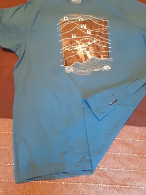 Blå t-shirt med grafiskt tryck Trespass - Cool blå t-shirt från Trespass med ett stort grafiskt tryck på bröstet som har topografiska linjer och text. T-shirten har rund hals och korta ärmar, perfekt för en avslappnad och modern look. Materialet är mjukt och bekvämt bomull. Unisex. Storlek M/L. Mått på bilderna. 