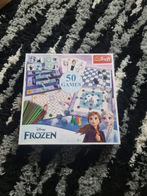 Disney Frozen 50 spel Trefl - Säljer ett spelset med 50 olika klassiska bräd- och kortspel med Disney Frozen-tema från Trefl. Setet innehåller spelplaner, spelpjäser, tärningar, kort och instruktioner, allt i en vit och lila box med motiv från Frozen. Perfekt för familjekvällar och barn från 5 år.