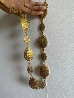 vintage Guldigt bälte med medaljongdetaljer - Unikt bälte i guld med stora ovala medaljonger och detaljerade mönster. Bältet har en bohemisk vibe och är perfekt för att lyfta vilken outfit som helst. Justerbar längd och metallkedja gör det enkelt att anpassa efter stil. 123cm length