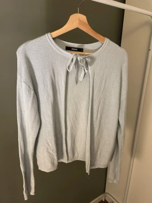 Ljusgrå stickad tröja med knyt - Ljusgrå stickad tröja från Bik Bok med rund halsringning och dekorativt knytband framtill. Tröjan har långa ärmar och är tillverkad i ett mjukt, ribbstickat material som ger en avslappnad och stilren look.