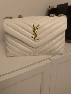 Vit Saint Laurent handväska i läder - Snygg vit handväska från Saint Laurent i quiltat läder med guldigt YSL-emblem framtill. Väskan har kedjerem och innerfack med dragkedja. Perfekt accessoar för att lyfta din outfit.
